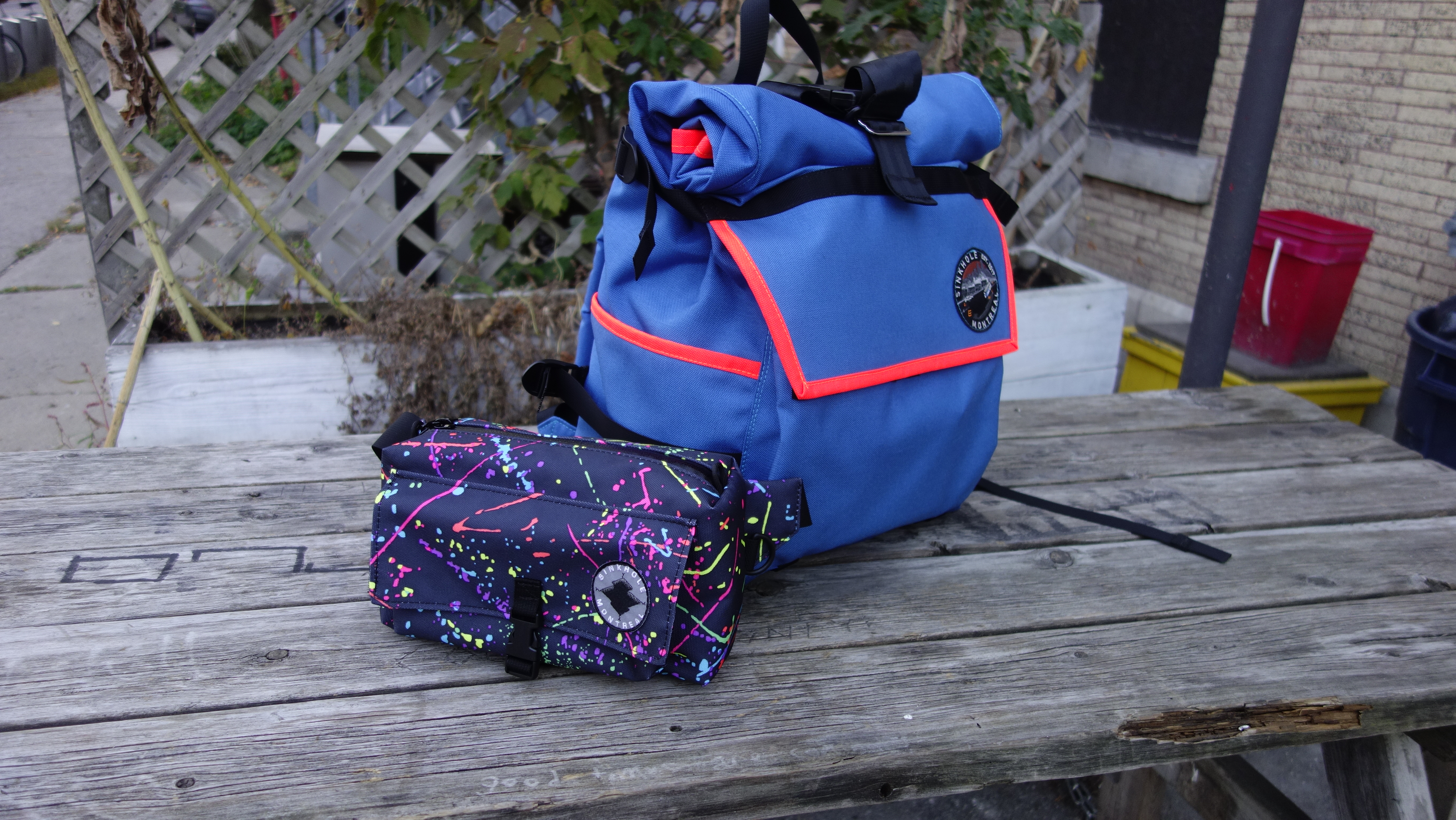 blue backpack with neon orange trim posed with an all-splatter fannypack on a picnic table